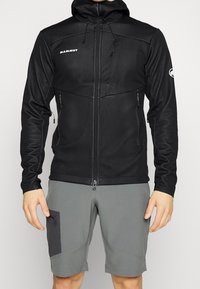Giacca softshell nera con zip frontale, cappuccio regolabile e numerose tasche, abbinata a pantaloni corti grigi con tasche laterali accentate.