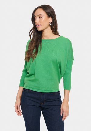 Maglione verde lavorato a maglia con scollatura ampia e maniche tre quarti, abbinato a jeans scuri. Il tessuto ha una trama delicata.