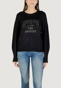 Schwarzer Pullover aus weichem Stoff, mit 'GUESS'-Logo und 'Los Angeles'-Text im metallischen Nieten-Design, langen Ärmeln und lockerer Passform.