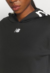 Czarna bluza z kapturem o gładkiej fakturze. Posiada biały logo oraz biały akcent wzdłuż kaptura, zaprojektowana do noszenia podczas aktywności sportowej.