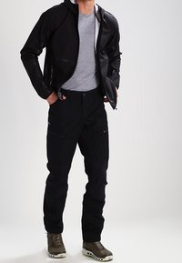 Veste noire imperméable avec fermeture éclair à l'avant et capuche, associée à un pantalon noir avec de nombreuses poches et des chaussures à semelles vertes. Texture lisse.