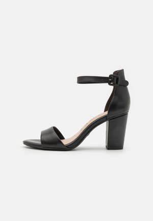 Sandale noire en cuir à bout ouvert avec talon bloc et bride de cheville avec boucle noire, vue de profil sur fond blanc.