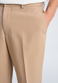 Beige skræddersyede bukser med sidelommer, båret af en person med den ene hånd afslappet placeret i lommen.