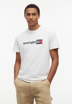 Egy fiatal férfi fehér "tommyjeans" feliratú pólóban és bézs nadrágban, zsebre tett kézzel áll egy sima fehér háttér előtt.