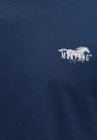 Logo cheval Mustang blanc avec texte sur tissu bleu foncé, centré sur la partie supérieure de la poitrine d'un vêtement.