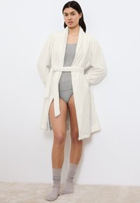 Accappatoio bianco in pile con colletto a scialle, cintura legata e spacchi laterali, indossato sopra una canotta grigia e pantaloni corti abbinati, abbinato a calzini a coste.