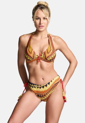 Blondynka w żółtym, pomarańczowym i czarnym wzorzystym bikini halter z frędzlami, pozująca na białym tle.