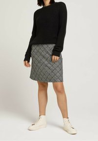 Mujer vestida con un suéter negro de punto, una falda gris de cuadros hasta la rodilla y zapatillas blancas de caña alta, de pie contra un fondo neutro.