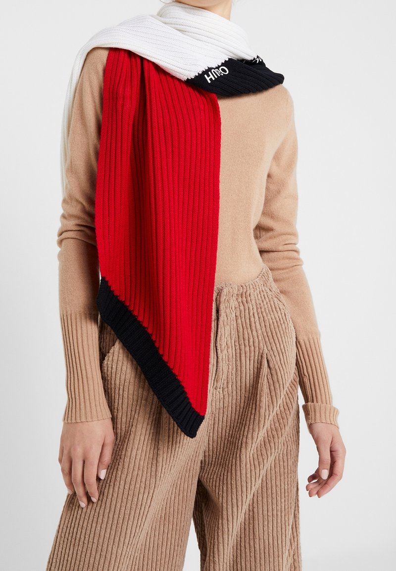Personne portant un pull côtelé beige et un pantalon large en velours côtelé, avec une écharpe en tricot color-block blanche, rouge et noire enroulée autour du cou.