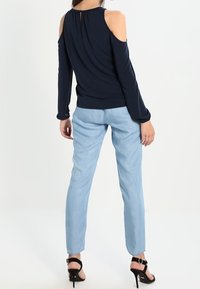 Tmavě modrý top s odhalenými rameny a volně splývajícím textilním detailem; světle modré úzké džíny; černé sandály na vysokém podpatku. Jednoduchý, čistý design bez viditelných vzorů.