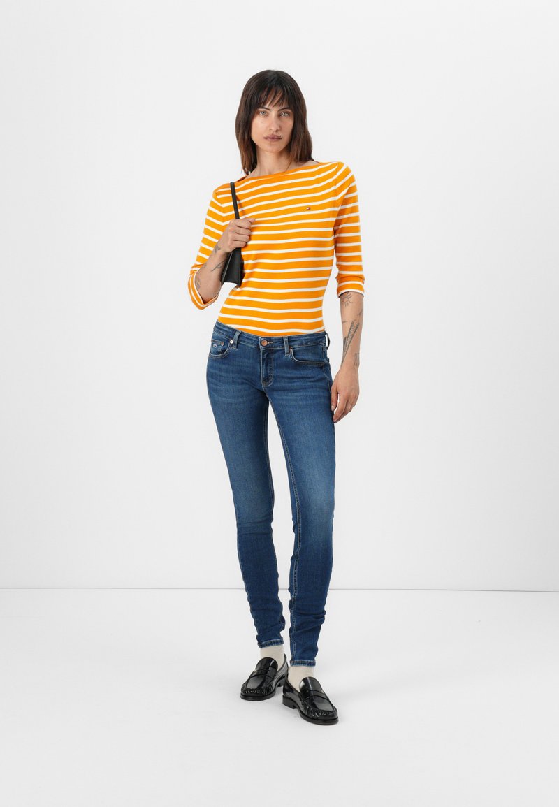 Pomarańczowa i biała w paski koszula z długim rękawem połączona z niebieskimi rurkami i czarnymi loafersami. Modelka trzyma małą czarną torbę.