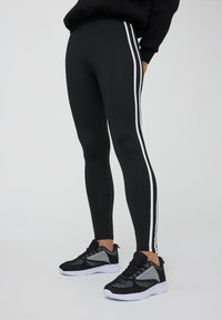 Leggings neri con strisce laterali bianche, realizzati in materiale elasticizzato. Abbinati a scarpe da ginnastica nere e grigie.