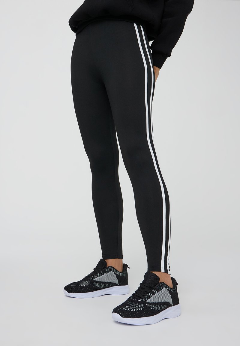 Leggings neri con strisce laterali bianche, realizzati in materiale elasticizzato. Abbinati a scarpe da ginnastica nere e grigie.