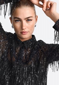 Bluză neagră din paiete cu un material transparent, detalii cu franjuri, guler și închidere cu nasturi. Design texturat și structurat.