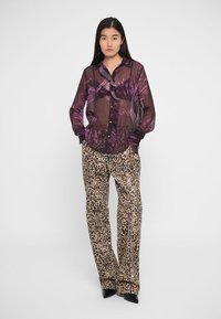 Blusa trasparente viola con motivo floreale, abbinata a pantaloni a stampa leopardata. La blusa ha una chiusura a bottoni; entrambi i pezzi presentano una vestibilità rilassata.