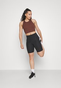 Smeđi sportski crop top i crne biciklističke kratke hlače visokog struka, s glatkom teksturom i bočnom džepom, u kombinaciji s crnim tenisicama i bijelim čarapama.