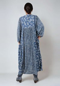 Lang blauwe jurk met bloemen- en paisleypatronen, geplooide achterkant, zachte stof en losse mouwen. Gestyled met grijze enkellaarzen.