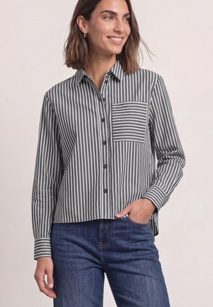 Femme portant une chemise boutonnée rayée verticale gris et blanc avec une poche poitrine à rayures horizontales et un jean bleu, souriante et regardant vers la droite.