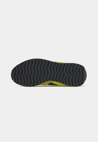 Potplat žutih tenisica s crnim cik-cak uzorkom i Reebok logom na potplatu, prikazan na jednostavnoj bijeloj pozadini.