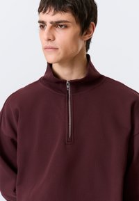 Sweatshirt de cor bordô com fecho éclair, gola alta, feito de tecido suave, apresentando um fecho prateado e design de corte descontraído.