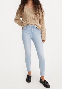 Suéter de punto beige con cuello en V, combinado con jeans ajustados azul claro y mocasines negros. Material texturizado y diseño ajustado.