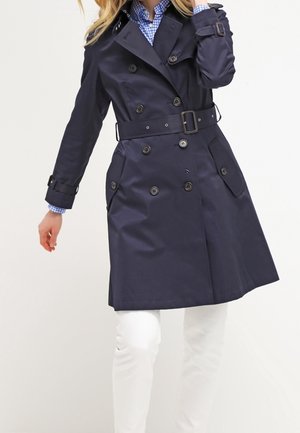 Vrouw draagt een marineblauwe double-breasted trenchcoat met riem, over een blauw geruit hemd en witte broek, staand tegen een effen achtergrond.