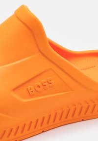 Gros plan sur une chaussure orange en caoutchouc à enfiler avec une semelle texturée et le logo "BOSS" embossé sur le panneau latéral.