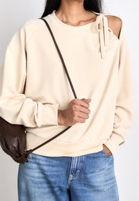 Beżowa bluza z odkrytymi ramionami wykonana z miękkiego materiału, z kokardą na jednym ramieniu. Połączona z jasnoniebieskimi jeansami.