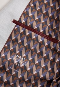 Geometrisch gemusterter Stoff in Braun, Blau und Beige mit burgunderfarbenem horizontalem Besatz und weißem Text mit der Aufschrift "NEXT ATHLETIC FIT 40R Made in Vietnam."