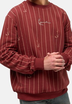 Mężczyzna w bordowej bluzie z okrągłym dekoltem, z cienkimi pionowymi beżowymi paskami i wyszywanym logo podpisu, z rękami złączonymi na przodzie.