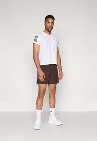 Λευκό αθλητικό πουκάμισο με μαύρες ρίγες στους ώμους, συνδυασμένο με καφέ σορτς που έχουν ανοιχτές διακριτικές ρίγες. Το σύνολο ολοκληρώνουν λευκά αθλητικά παπούτσια και κάλτσες.