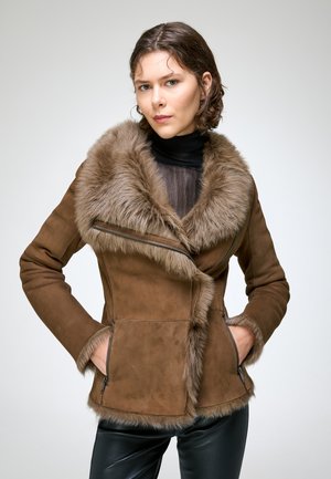 Femme aux cheveux courts et bruns portant une veste en daim marron doublée de fourrure et un col roulé noir, debout les mains dans les poches devant un fond uni.