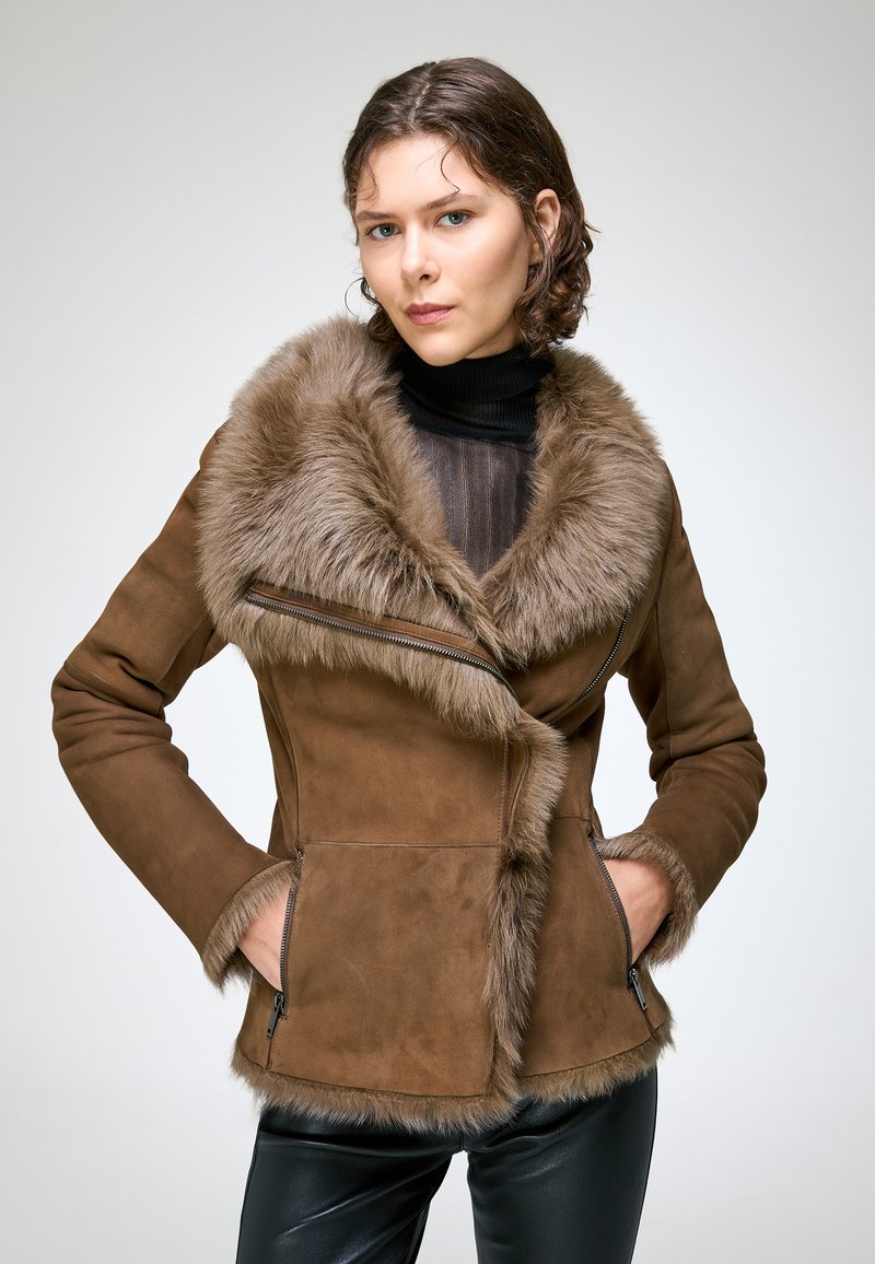 Femme aux cheveux courts et bruns portant une veste en daim marron doublée de fourrure et un col roulé noir, debout les mains dans les poches devant un fond uni.