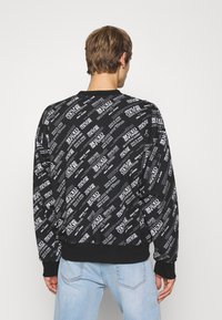 Černá mikina s opakujícím se bílým textovým vzorem. Dlouhé rukávy s žebrovanými manžetami a lemem. Měkká textura, volný střih. Zobrazeno zezadu.