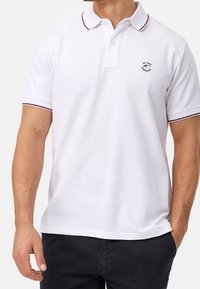 Chemise polo blanche avec un plastron à deux boutons, manches courtes et une fine bordure bourgogne autour du col et des manches. Présente un petit logo sur la poitrine.