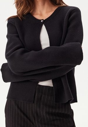 Femme portant un cardigan noir boutonné sur un haut blanc avec un pantalon noir à fines rayures, bras croisés sur la poitrine.