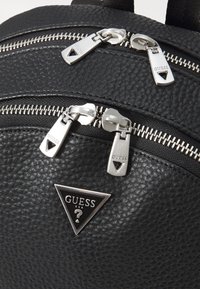 Un sac à dos noir texturé présentant trois compartiments zippés et des accessoires métalliques. Décoré d'un logo Guess en forme de triangle.