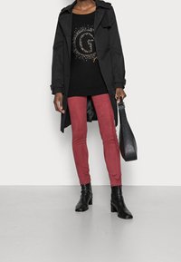 Schwarzer Trenchcoat über einem schwarzen Pullover mit verziertem Buchstaben "G", kombiniert mit weinroten Skinny-Jeans und schwarzen Ankle Boots. Schwarze Handtasche.