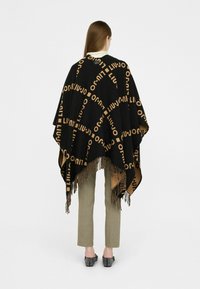 Zwarte poncho met een tan logo patroon, met Franje aan de onderkant, gedragen over beige broeken. Model kijkt weg, waardoor het ontwerp en de textuur te zien zijn.