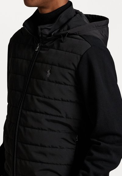 Μαύρο επενδεδυμένο μπουφάν με φερμουάρ και κουκούλα. Διαθέτει σχεδίαση καπιτονέ και μαύρες μεταλλικές λεπτομέρειες. Λογότυπο στο στήθος, υφασμάτινα μανίκια.