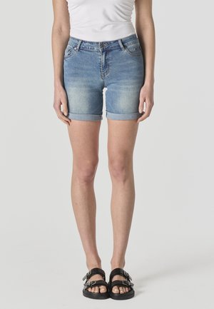 Mujer con shorts de mezclilla azul claro con dobladillo enrollado, top blanco sin mangas y sandalias negras con hebilla, de pie contra un fondo liso.