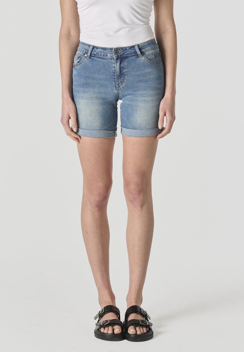 Žena v svetlomodrých rifľových šortkách s vyhrnutým lemom, bielom bezrukávovom topu a čiernych sandáloch so sponou stojaca pred jednoduchým pozadím.