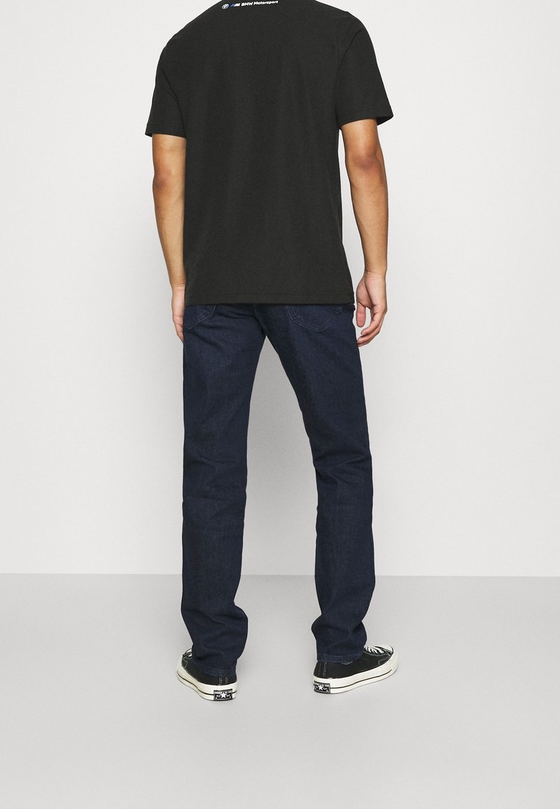 Homme se tenant de dos, portant un t-shirt noir de BMW Motorsport, un jean bleu foncé et des baskets noires et blanches.