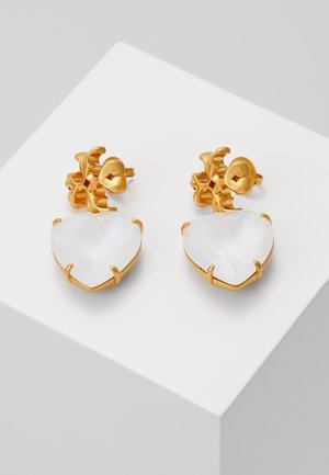 Złote kolczyki z białymi kamieniami w kształcie serca i misternym złotym zdobieniem na górze, prezentowane na białej, pochylonej powierzchni.