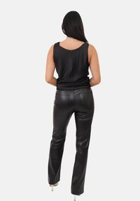 Donna con lunghi capelli scuri che indossa un top senza maniche in satin nero, pantaloni di pelle neri e scarpe con tacco beige, in piedi con le spalle rivolte verso la macchina fotografica.