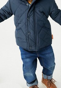 Námořnická prošívaná bunda s patentkami a předními kapsami, zkombinovaná s modrými džíny, ohrnutými na manžetách, a hnědými šněrovacími botami.