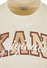 Cream-colored sweatshirt featuring bold brown and light tan lettering spelling "KAN" and "Original." Made of soft fabric with a crew neck design.