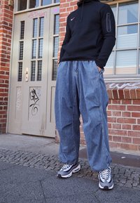 Felpa nera con cappuccio e tasca con zip, abbinata a pantaloni in denim azzurro chiaro larghi e sneakers grigie con dettagli blu. Sfondo di muro di mattoni.