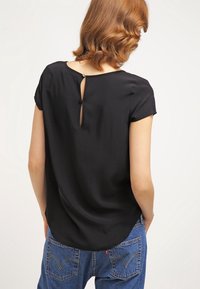 Czarna bluzka z krótkim rękawem z detalem w kształcie dziurki z tyłu. Miękki materiał o gładkiej strukturze, zestawiona z niebieskimi dżinsami.