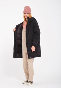 Zwarte pufferjack met ritssluiting aan de voorkant, zijzakken en een capuchon, gedragen over beige corduroy overall en een roze shirt, met een gebreide beanie.